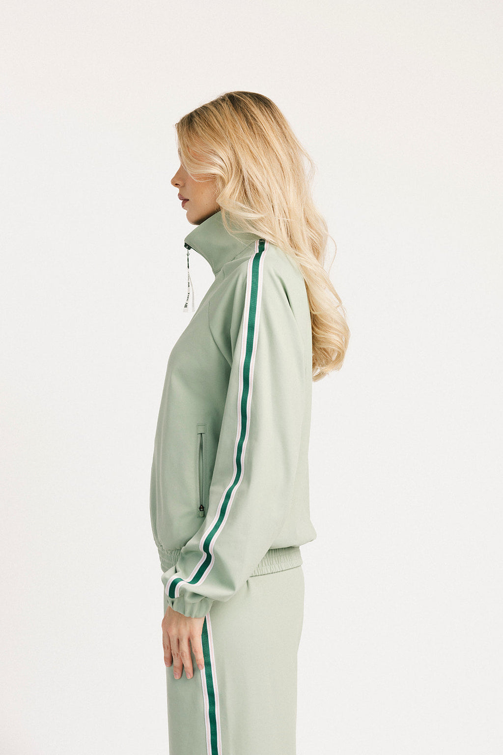 Woman wearing a light green jacket with a high collar and white pants on a white background